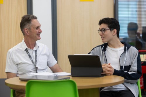 male student and male teacher sitting at a desk and chatting