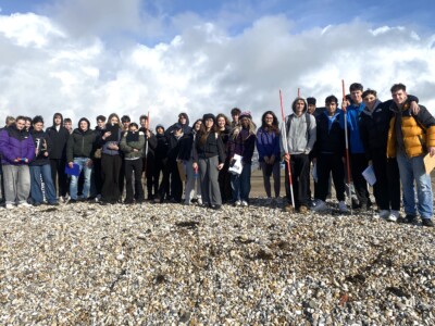 A large group of geography students standing on the pebbles at the beach wearing coats