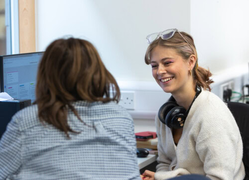 Two female students smiling and chatting to each other in front of computers