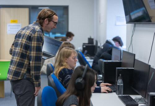 A male learning support teacher assisting students at the computers in the study centre