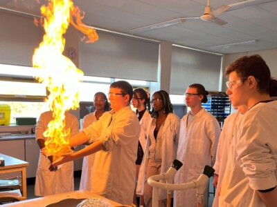 A group of chemistry students in lab coats observing a student with flames coming out of his hands