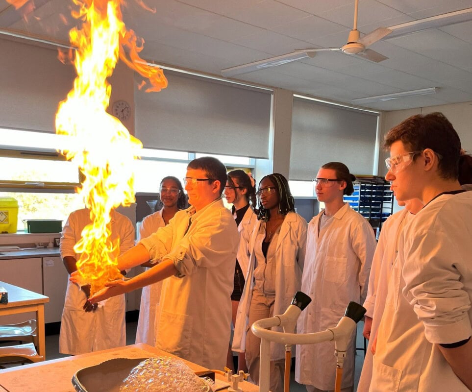 A group of chemistry students in lab coats observing a student with flames coming out of his hands