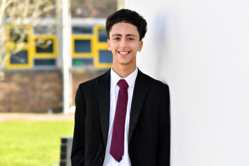 portrait of a student in a smart suit and red tie smiling as he leans against a white wall