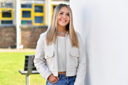 student wearing a white jacket and jeans poses against a white wall